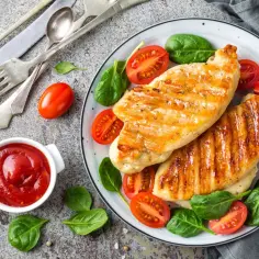 Grillowana pierś kurczaka z pomidorkami koktajlowymi i szpinakiem na talerzu, obok miseczka ketchupu.